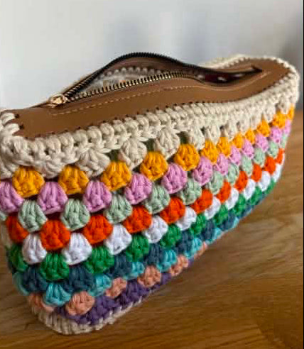 A handmade crocheted zipper pouch with a tan leather-look top trim and a multicolor striped crochet body, shown on a wooden surface against a light wall.