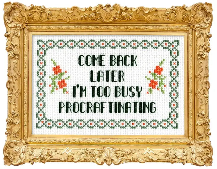 Framed cross-stitch style wall art with an ornate gold frame. The stitched design shows black text reading “COME BACK LATER I’M TOO BUSY PROCRAFTINATING” with green and red floral motifs and a decorative border on a white fabric background.