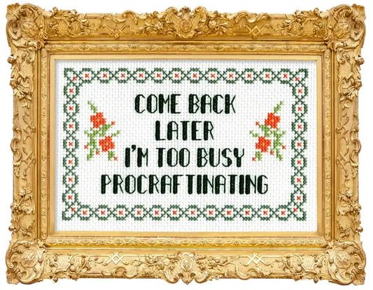 Framed cross-stitch style wall art with an ornate gold frame. The stitched design shows black text reading “COME BACK LATER I’M TOO BUSY PROCRAFTINATING” with green and red floral motifs and a decorative border on a white fabric background.